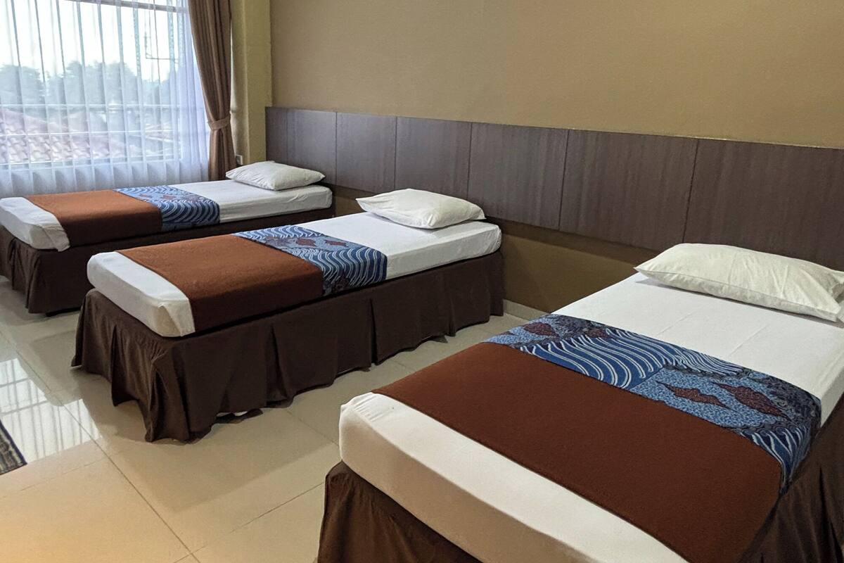 A hotel room with three single beds, each with a brown skirted base and white linens. The room has a beige wall and a window with curtains.