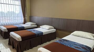 Habitación de hotel con tres camas individuales, cada una con falda marrón y ropa de cama blanca. La pared es color beige y hay una ventana con cortinas.
