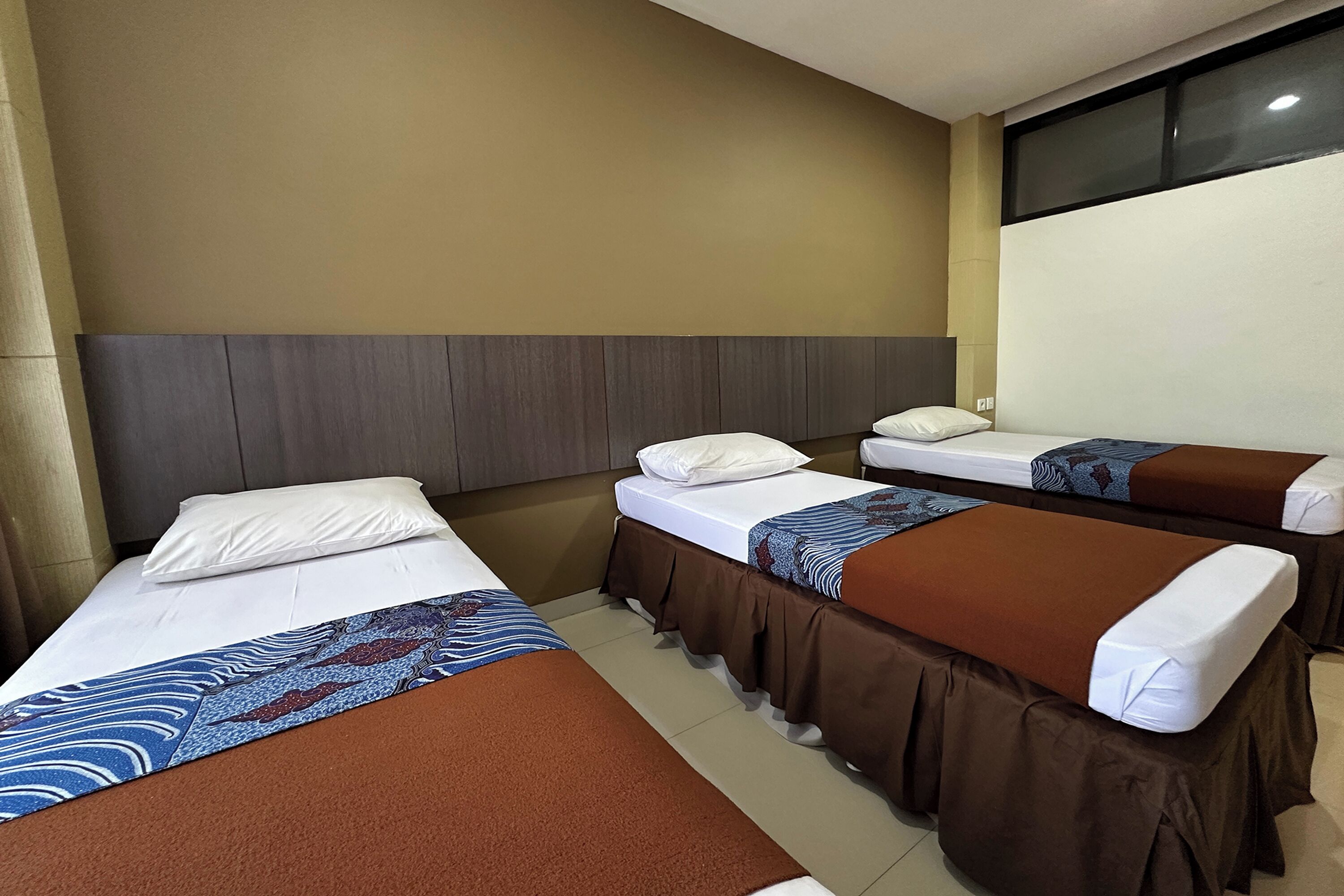A hotel room with three single beds, each with a brown bedspread and white pillows. The room has a neutral color scheme with beige walls and a dark wood headboard.