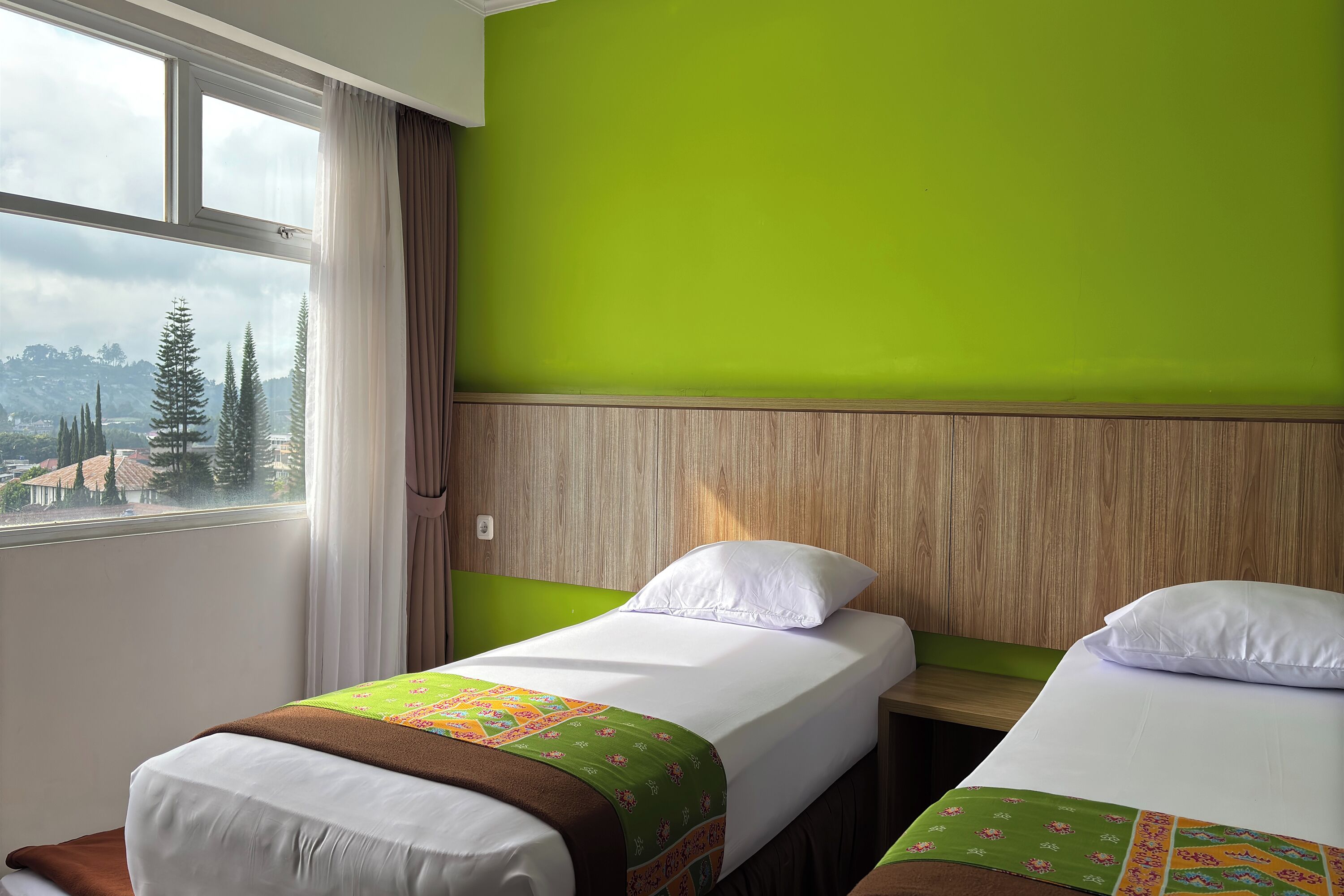 A hotel room with two beds, a large window with a view of trees and buildings, and a green wall.