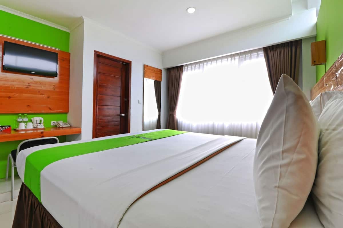 A hotel room with a bed, a desk with a telephone, a television mounted on a wooden panel, and a window with curtains.