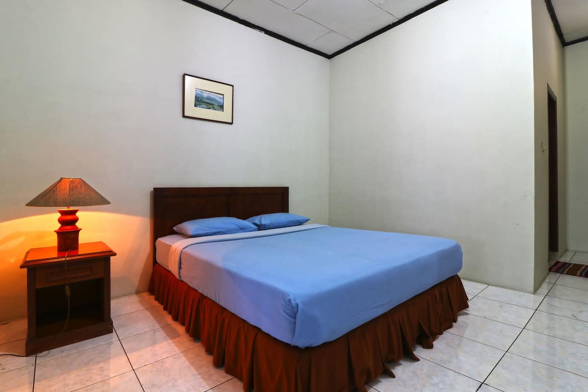 A neatly made bed with a blue bedspread and matching pillows, a wooden nightstand with a lamp, and a framed picture on the wall.