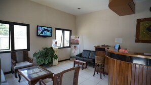 Um hall de hotel com balcão de recepção, área de estar, televisão e uma planta grande.