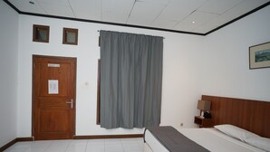 Quarto de hotel com porta de madeira, placa de check-out, cortina cinza, cama com cobertor cinza, mesa de cabeceira com luminária e quadro na parede.