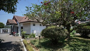 Uma casa com telhado de cerâmica, rodeada por um jardim bem cuidado com diversas plantas e uma pequena árvore com flores vermelhas.