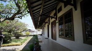 Uma galeria coberta com piso de azulejos, grandes janelas e área de jardim com diversas plantas.