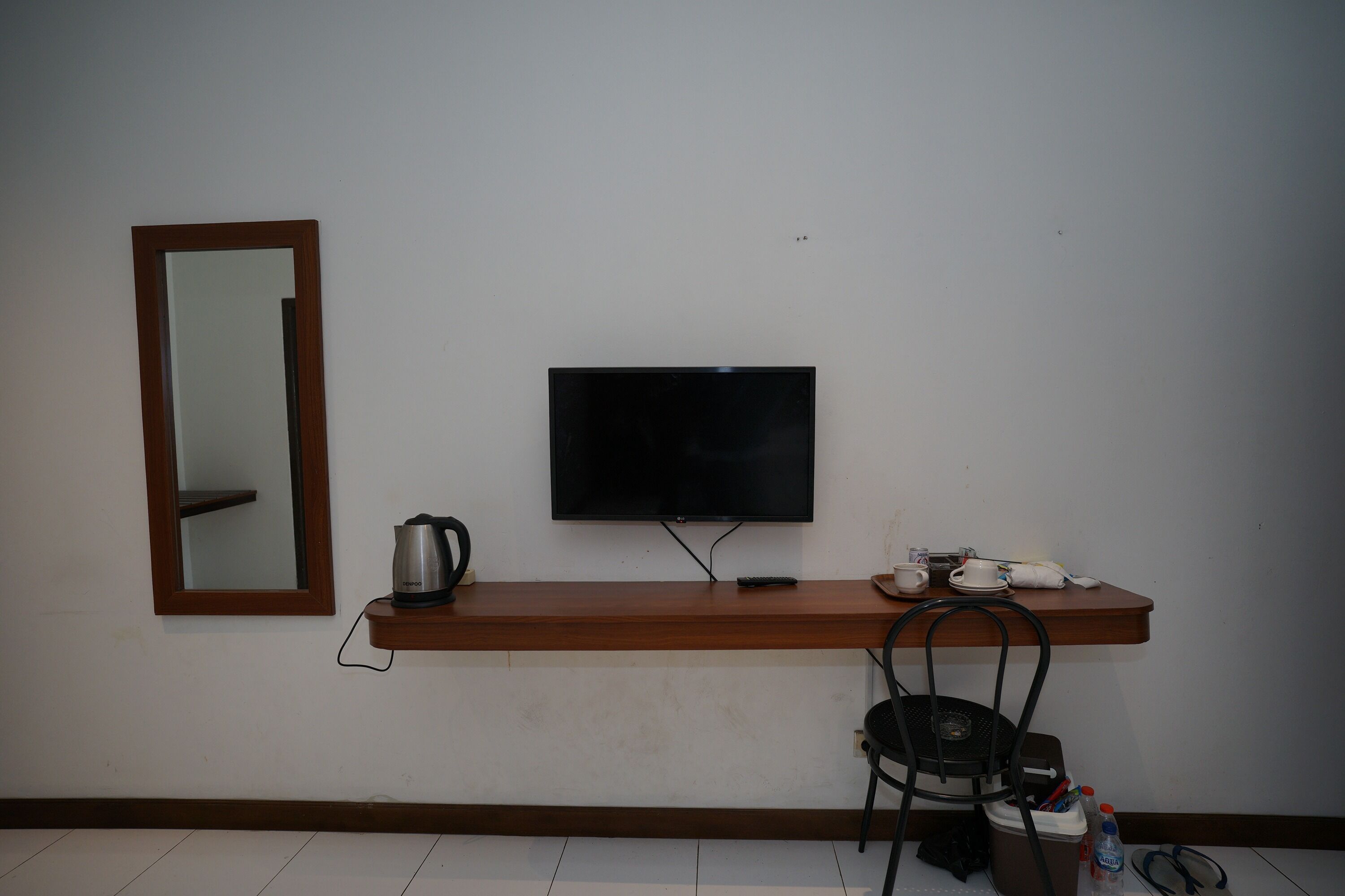 A hotel room with a wooden console table, a flat-screen TV mounted on the wall, a kettle, a remote control, a chair, and a small trash bin.