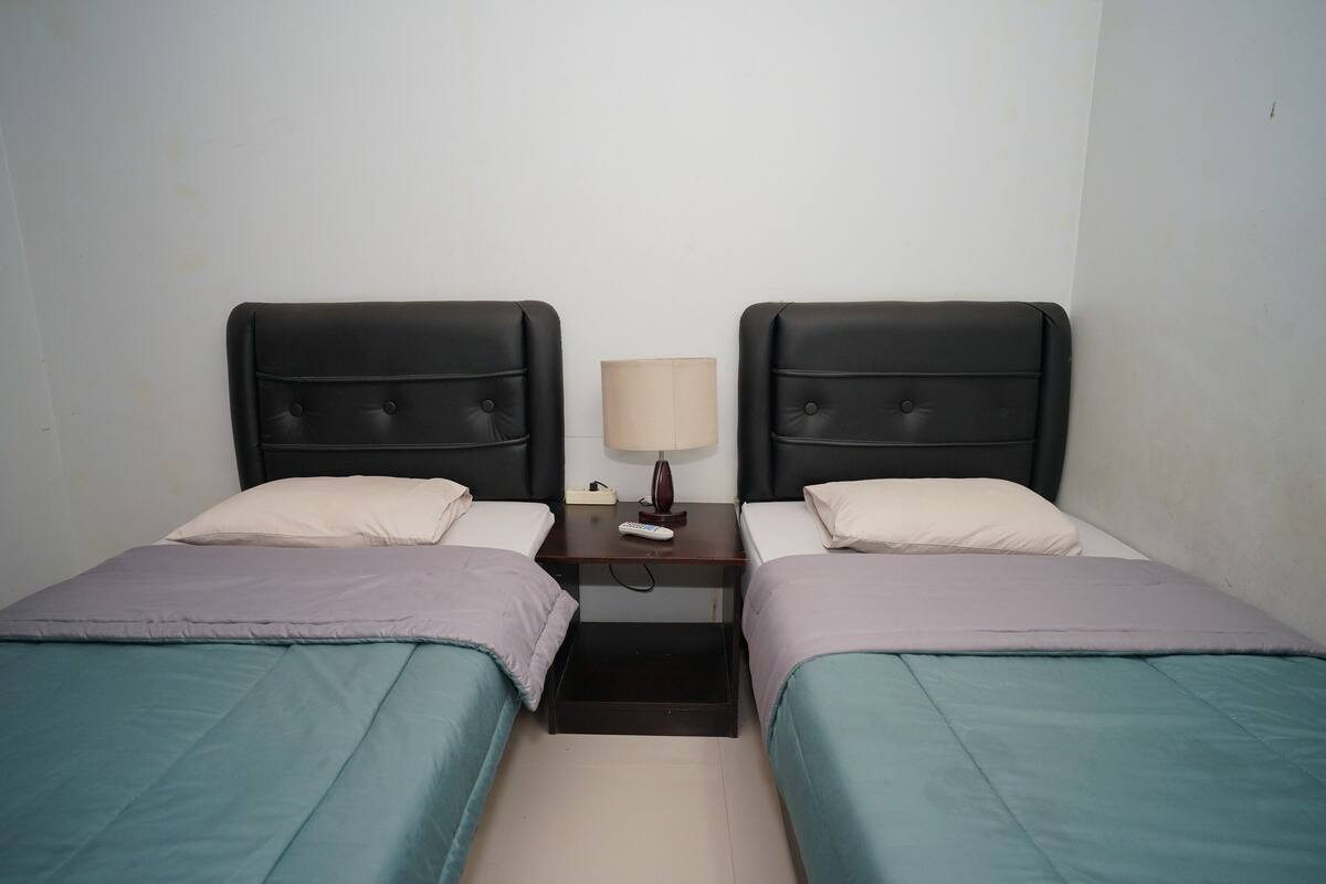 Habitación doble con dos camas, cada una con cabecera, mesita de noche con lámpara y un cajón pequeño.