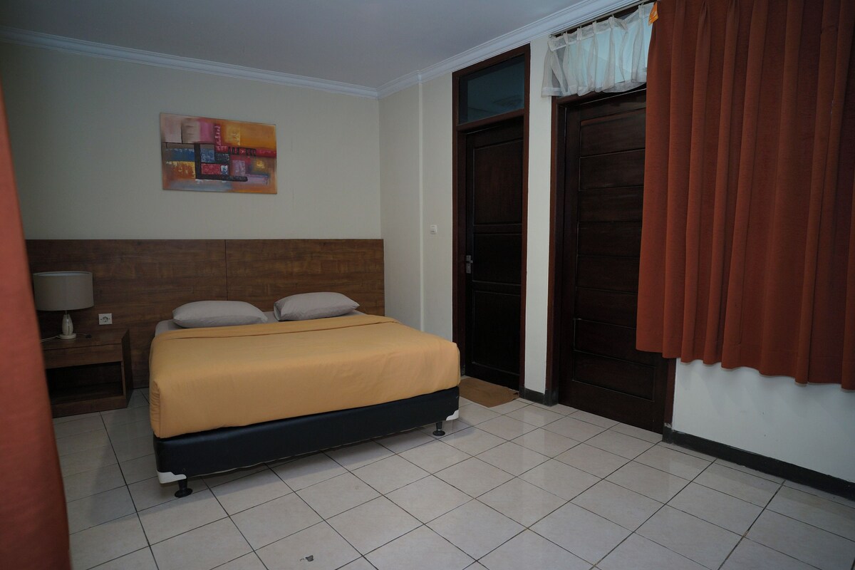 A hotel room with a bed, a nightstand, a painting on the wall, and a door with curtains.