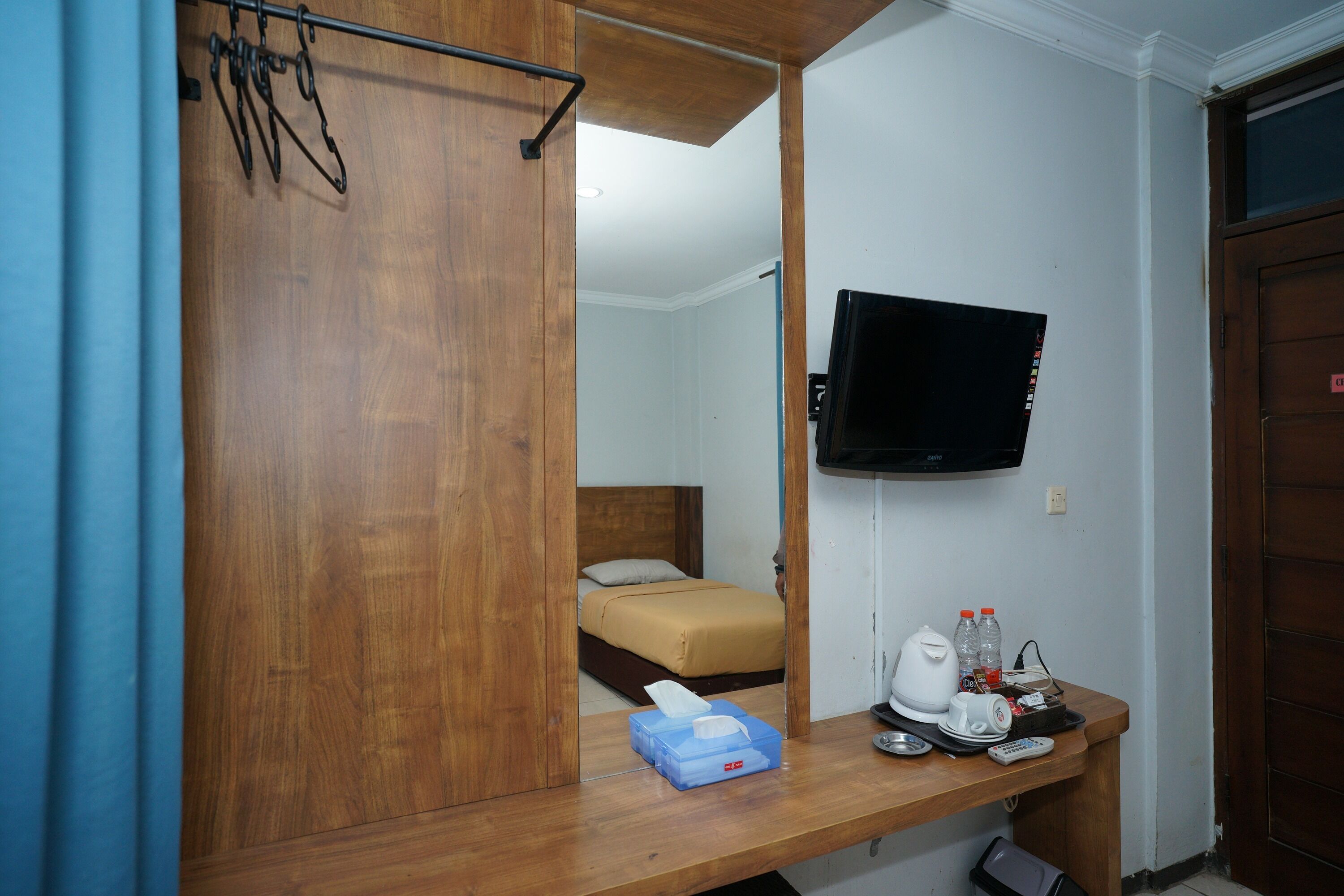 A hotel room with a wooden wardrobe, a bed with a white mattress, a television mounted on the wall, and a desk with a coffee maker, water bottles, and a remote control.