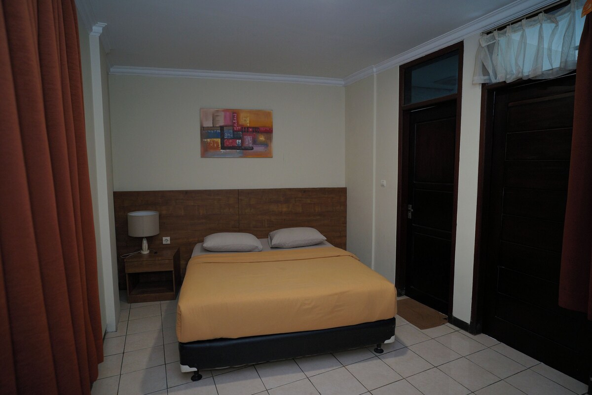A hotel room with a bed, a nightstand with a lamp, a painting on the wall, and a door.