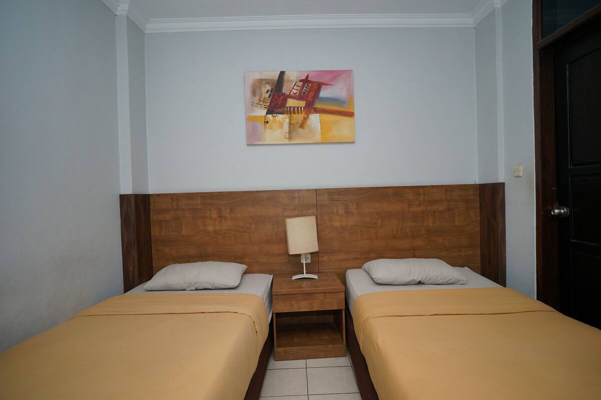 Una habitación de hotel con dos camas individuales, un cabecero de madera, una mesita de noche con una lámpara y un cuadro en la pared.