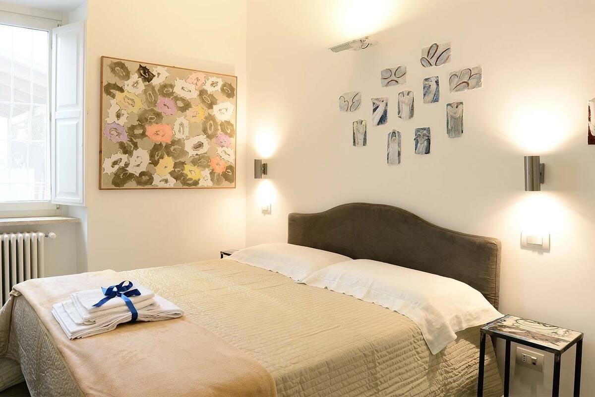 A neatly made bed with a folded towel and a blanket, a large abstract painting on the wall, and a series of framed artworks.