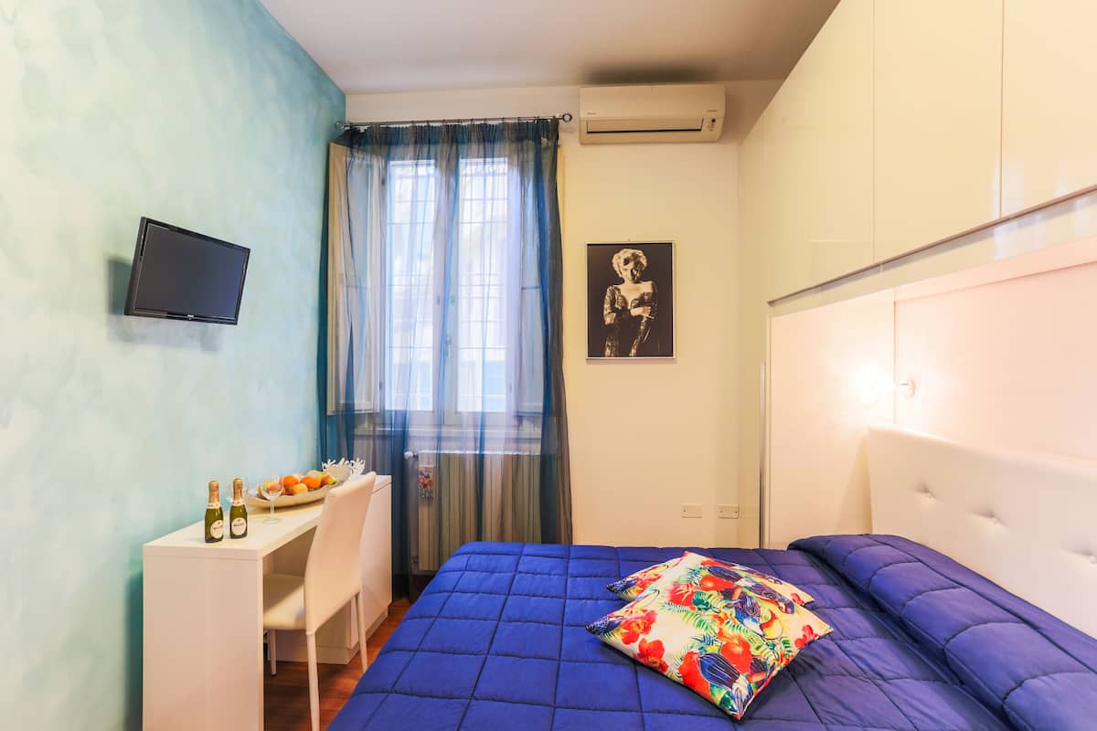 Ein Schlafzimmer mit Bett, Schreibtisch, Stuhl, Fernseher und einem Fenster mit Vorhängen.