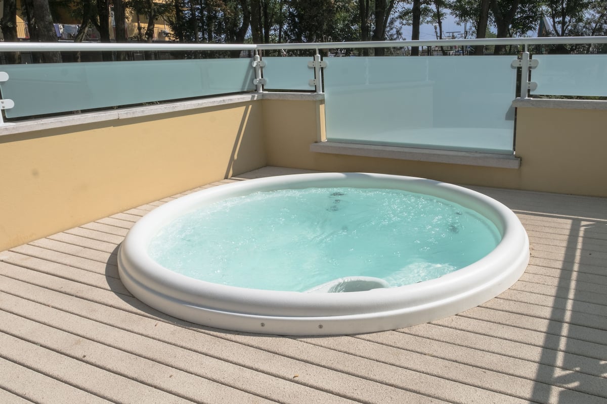 Ein Whirlpool auf einer gefliesten Terrasse mit Glasgeländer und einer beigen Wand im Hintergrund.