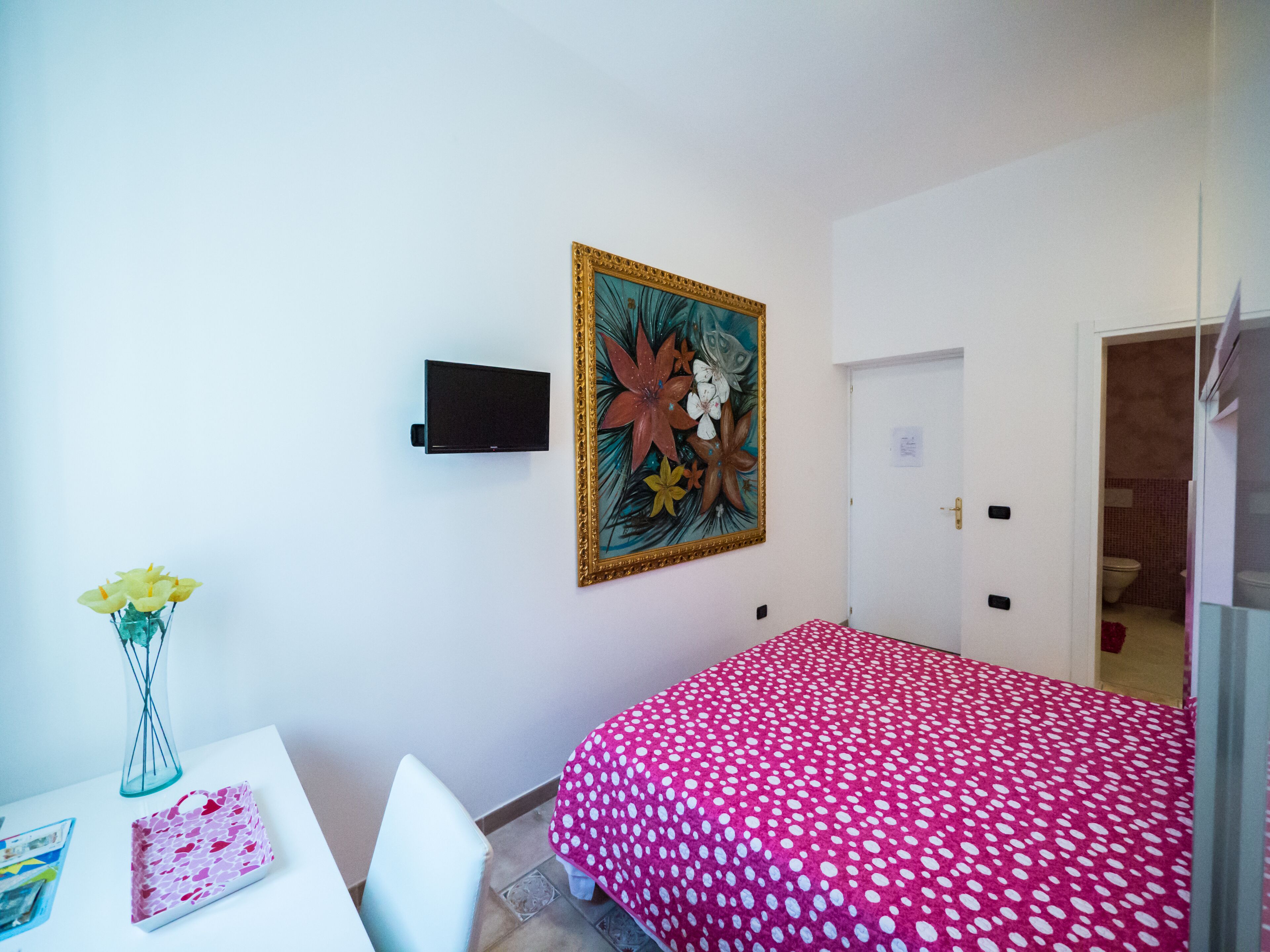 A bedroom with a bed, a TV mounted on the wall, a painting, and a small table with a vase.