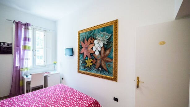 A bedroom with a bed, a desk, a chair, a window with curtains, and a wall-mounted painting. - Villa Bellini (Comacchio)