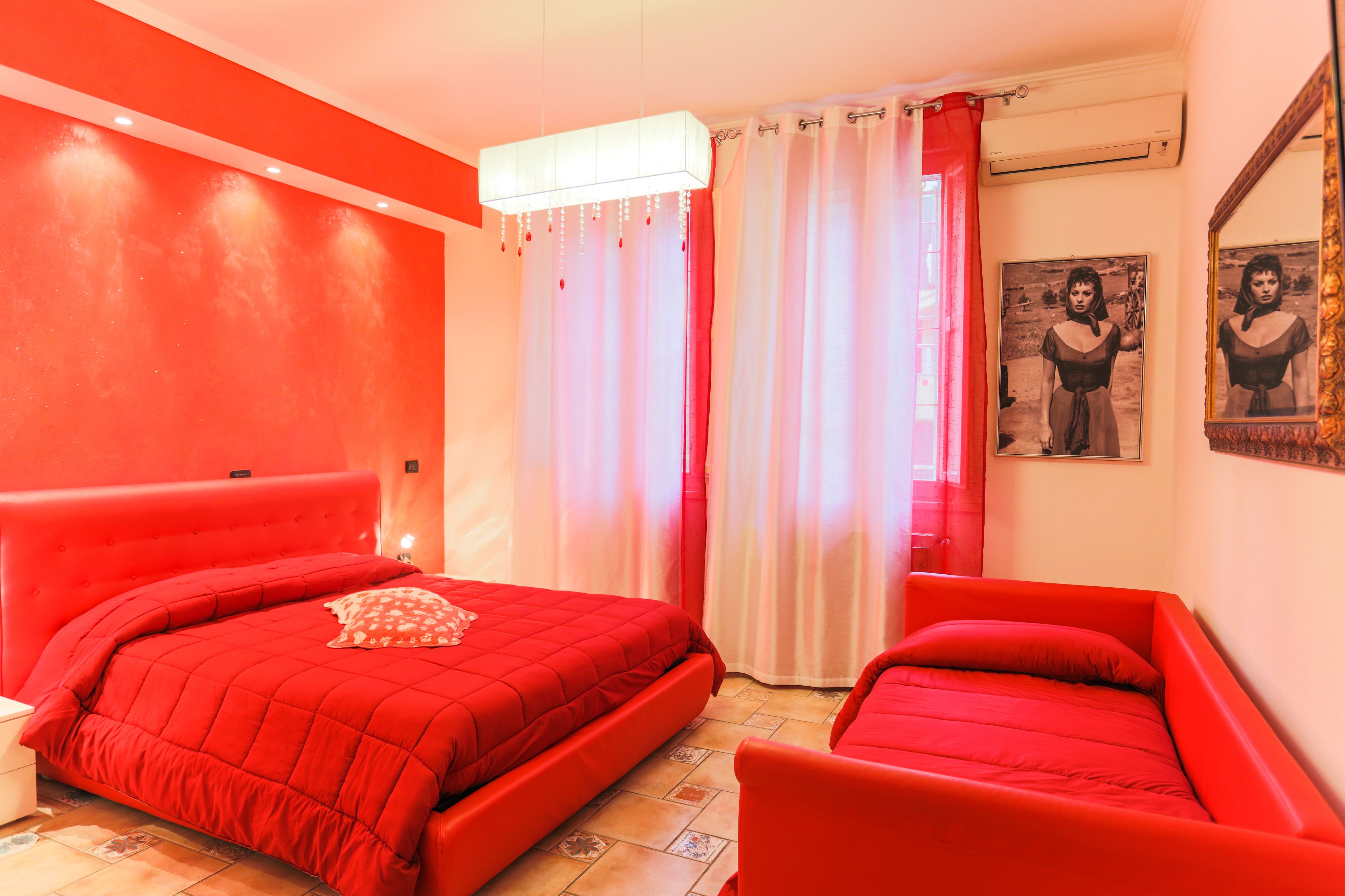 A room with a red bed, a smaller red chair, a nightstand, and a mirror on the wall.