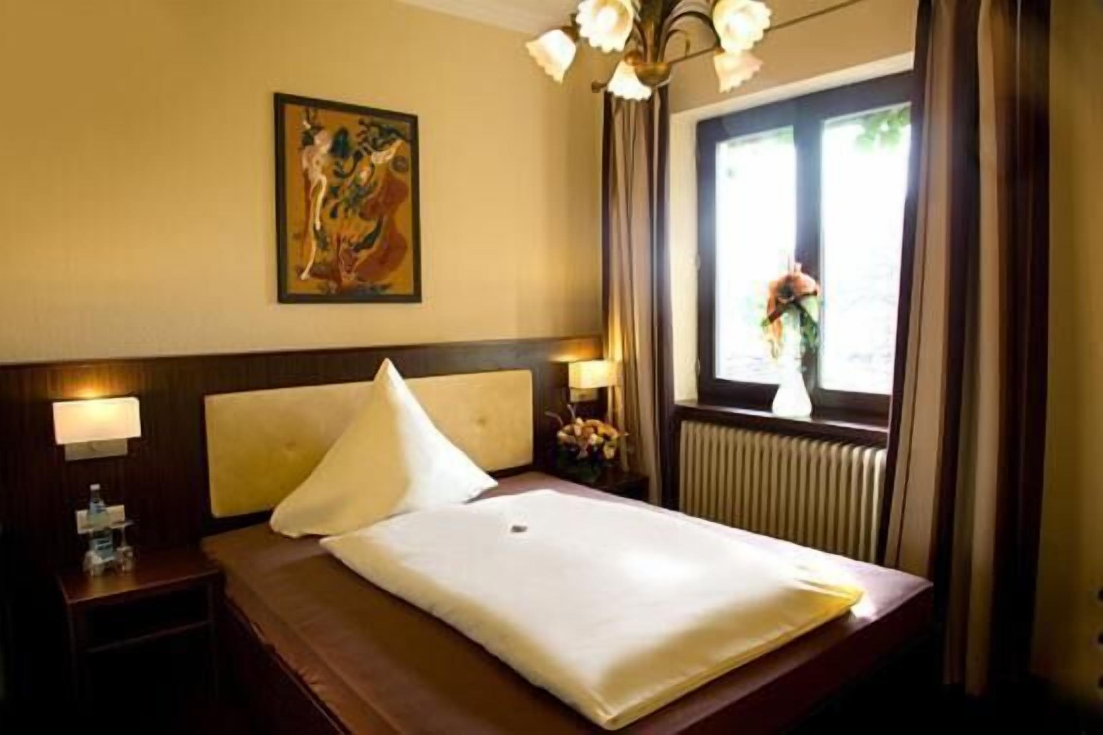 A hotel room with a bed, a nightstand, a lamp, a painting on the wall, and a window with curtains.
