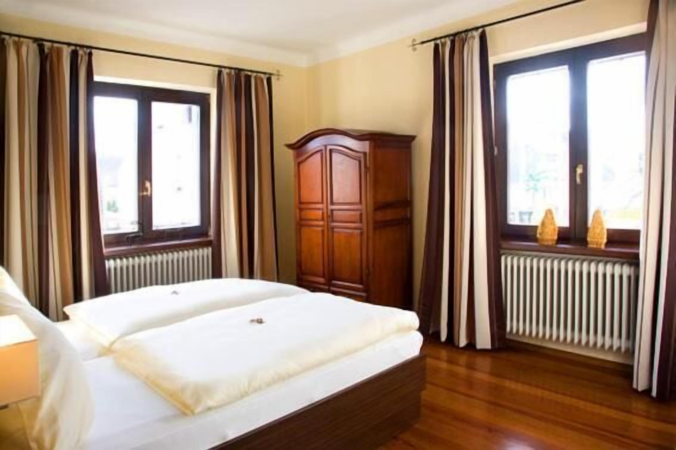 Quarto de hotel com cama de casal, guarda-roupa de madeira e duas janelas com cortinas.