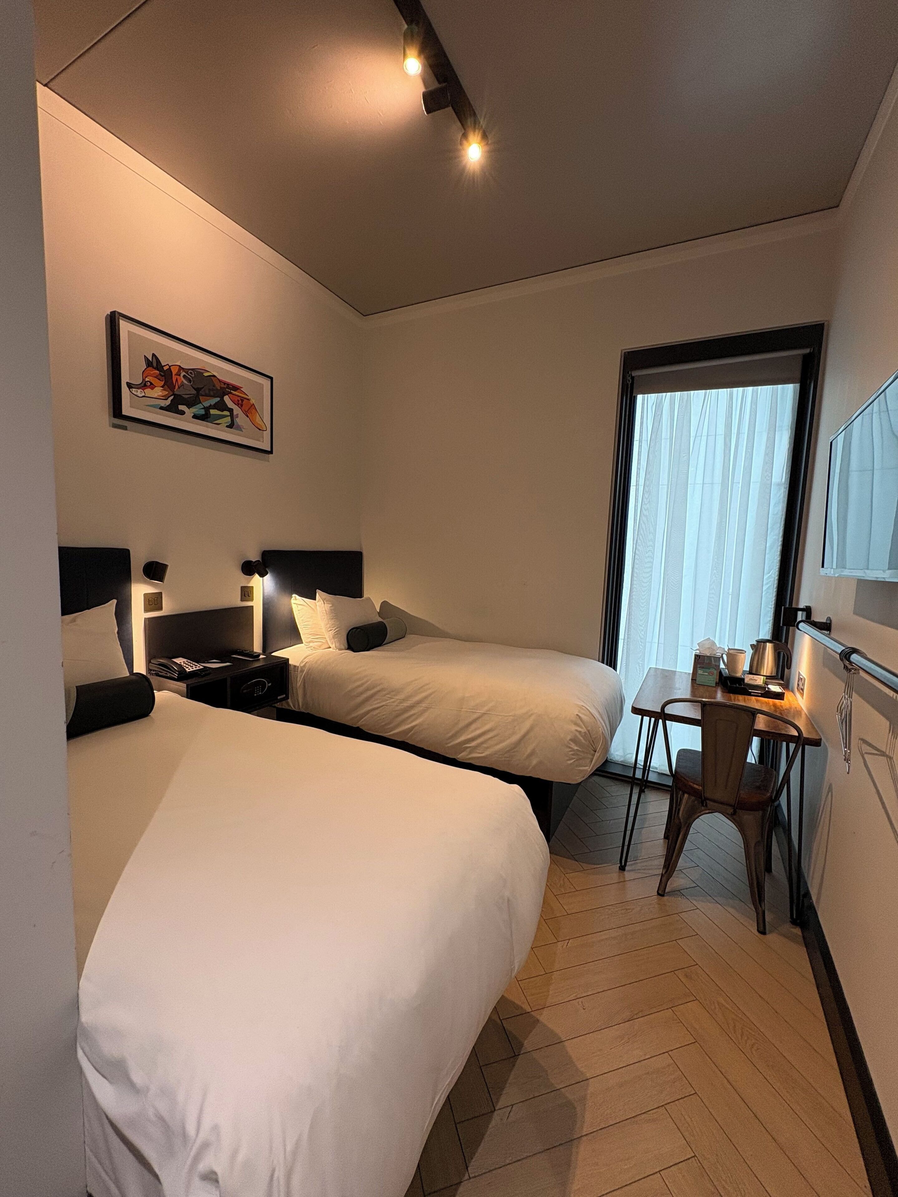 A hotel room with two beds, a desk, a chair, a window with curtains, a painting on the wall, and a ceiling light fixture.