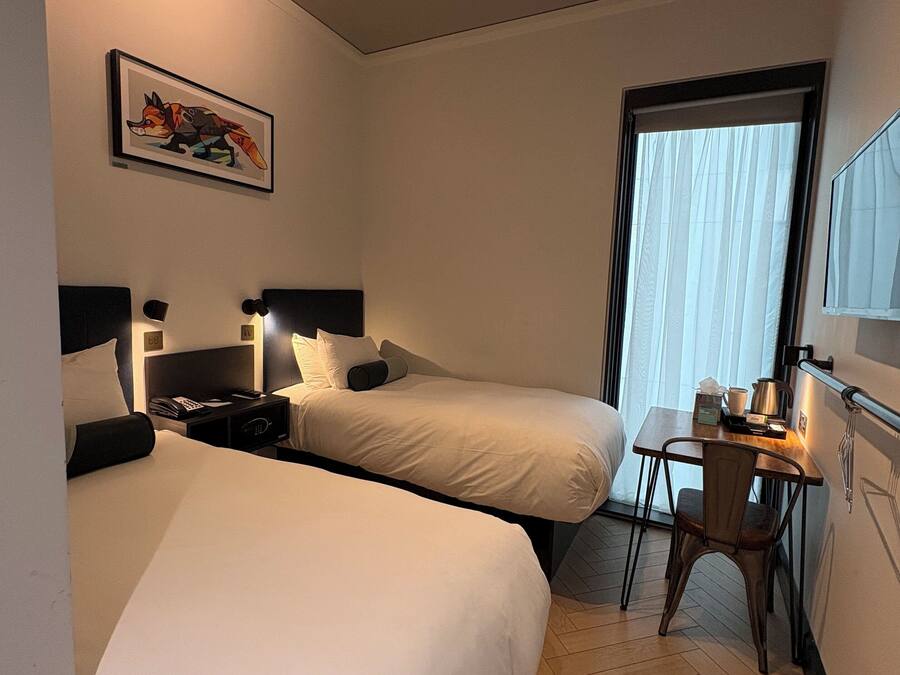A hotel room with two beds, a desk, a chair, a window with curtains, a painting on the wall, and a ceiling light fixture.