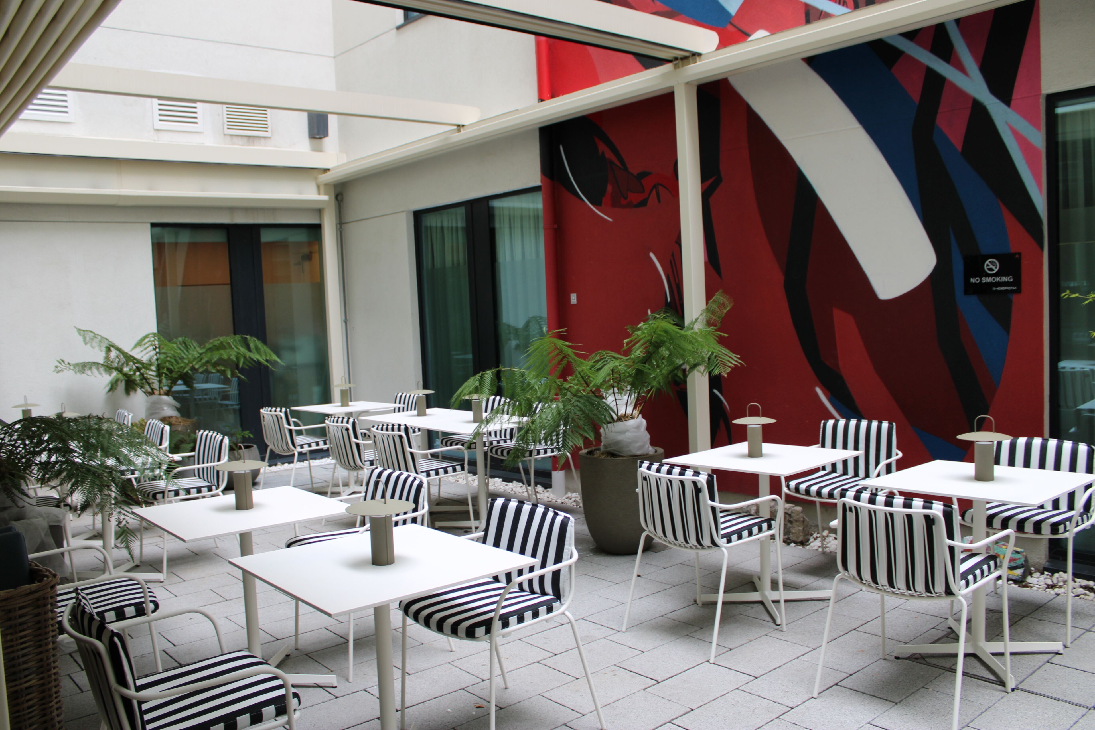 Un café al aire libre con mesas blancas y sillas a rayas, plantas en macetas y una pared roja con arte abstracto.
