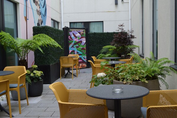 An outdoor seating area with wicker chairs and tables, surrounded by potted plants and a colorful mural on the wall.