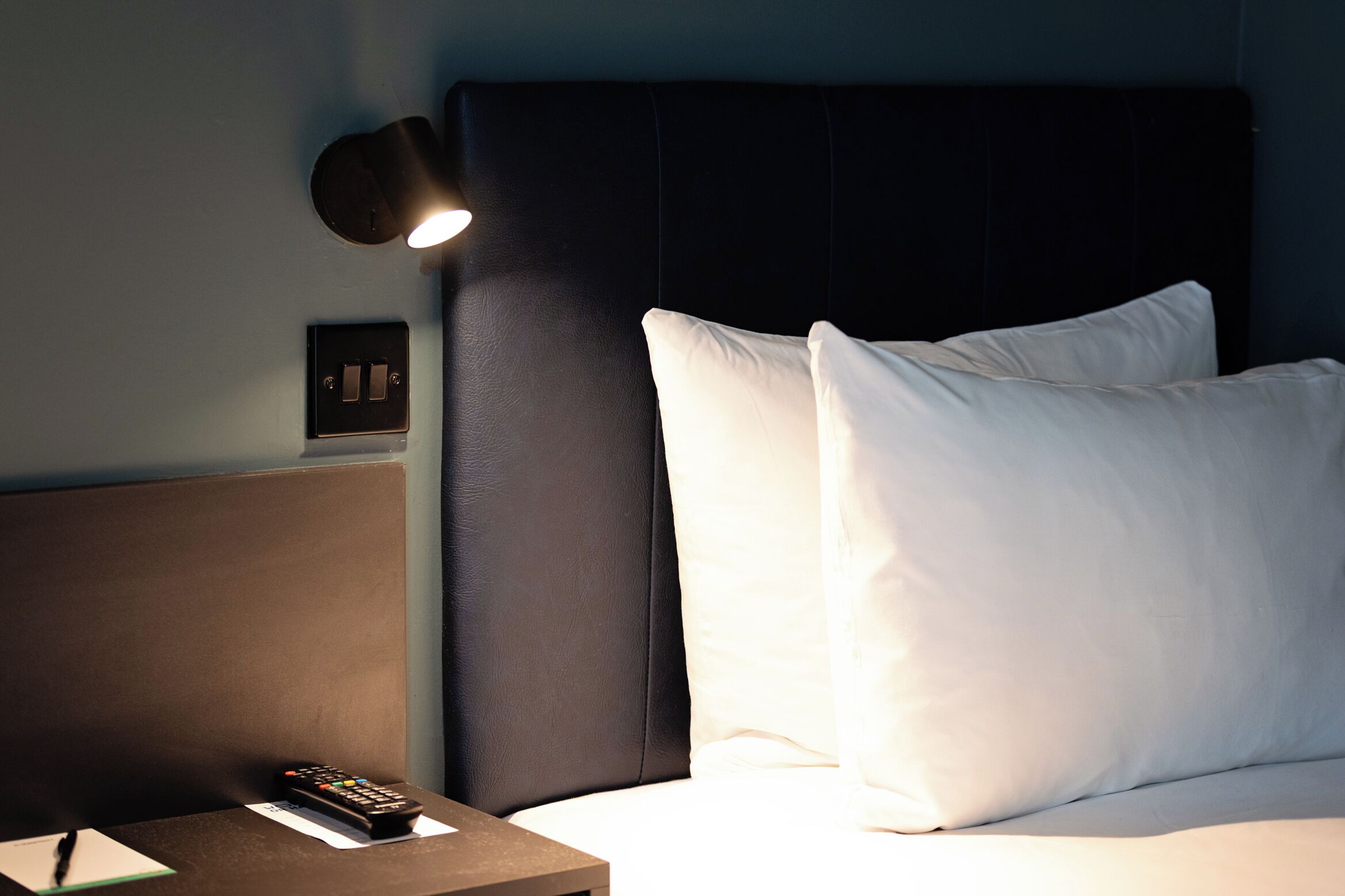 A hotel room with a bed, a bedside table with a remote control, a lamp, and a power outlet.