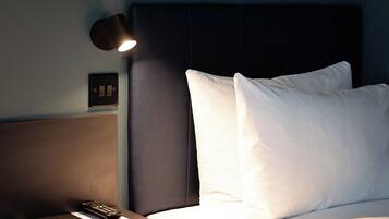 A hotel room with a bed, a bedside table with a remote control, a lamp, and a power outlet.