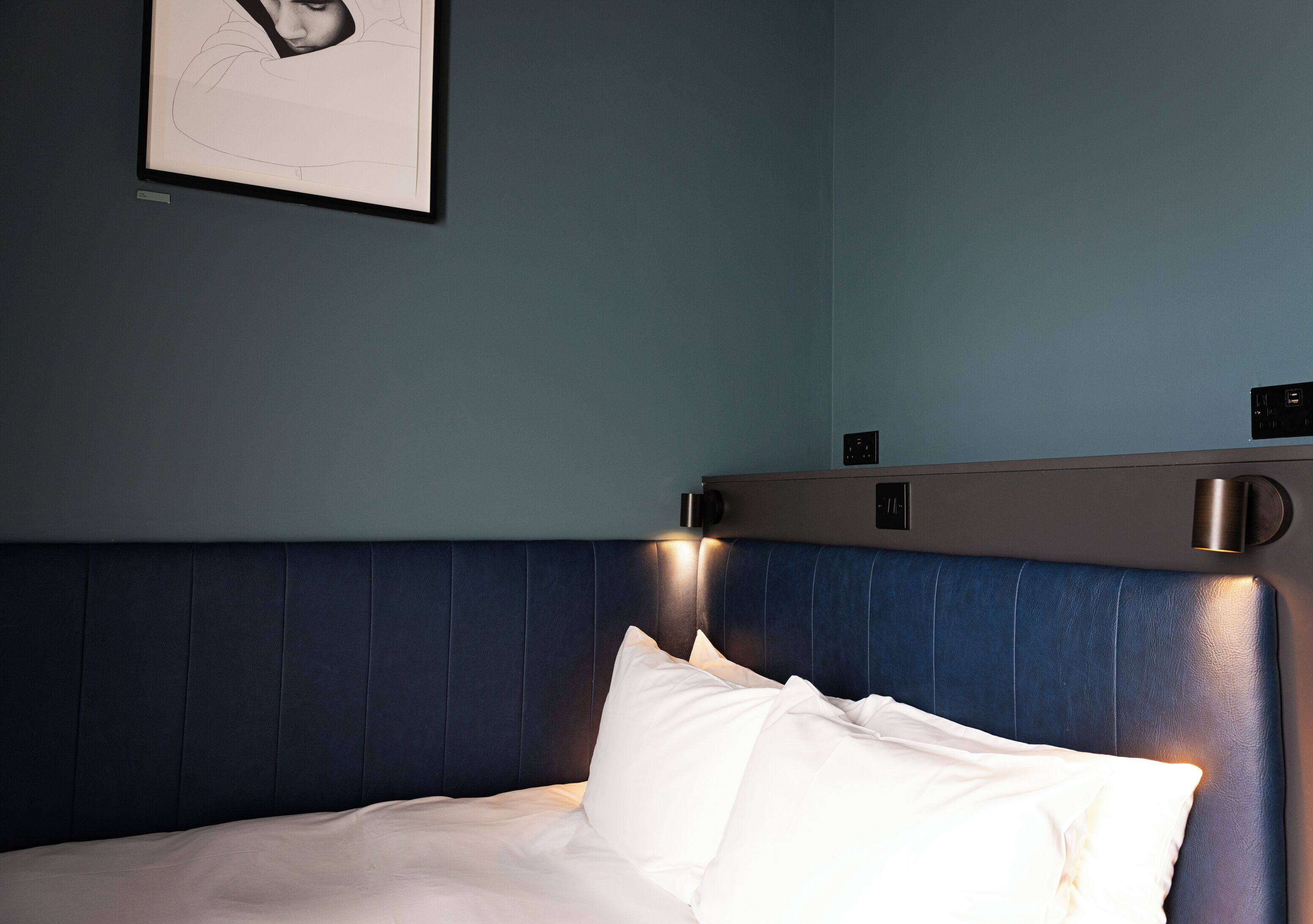 Een keurig opgemaakt bed met witte lakens, een donkerblauwe hoofdbord en een ingelijste foto aan de muur.
