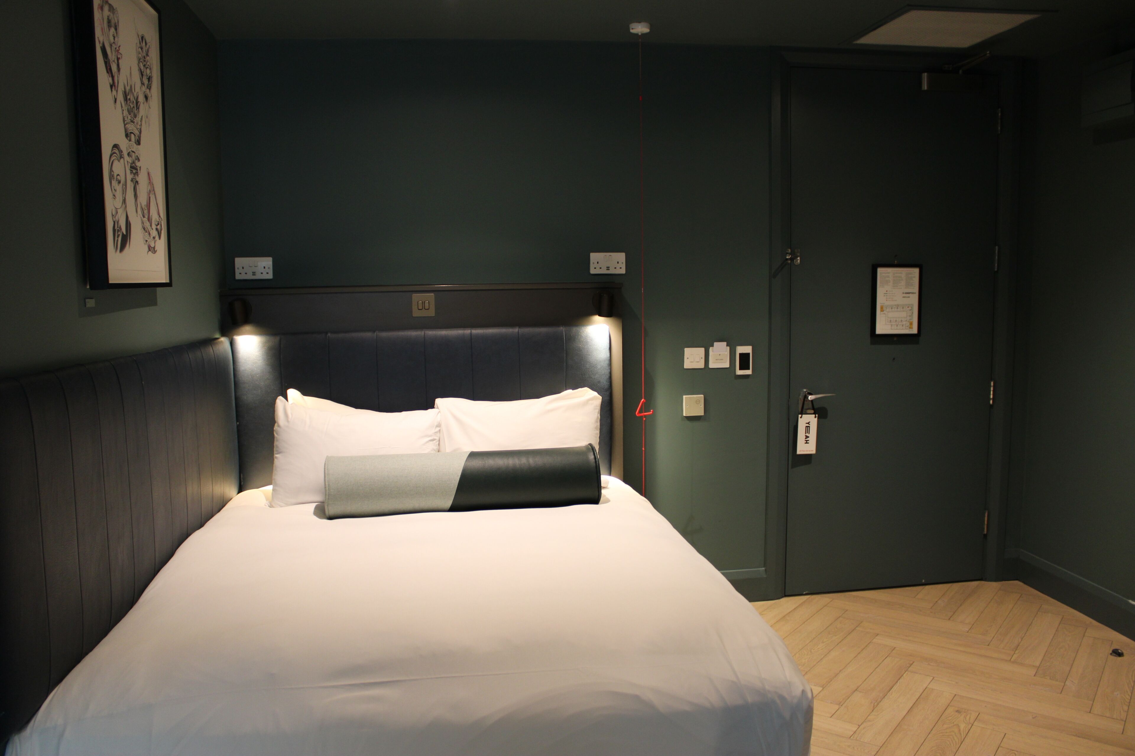 Un lit bien fait, avec des draps blancs et un plaid foncé, un luminaire fixé au mur au-dessus du lit, et une porte dotée d’une serrure et d’un porte-clés suspendu.
