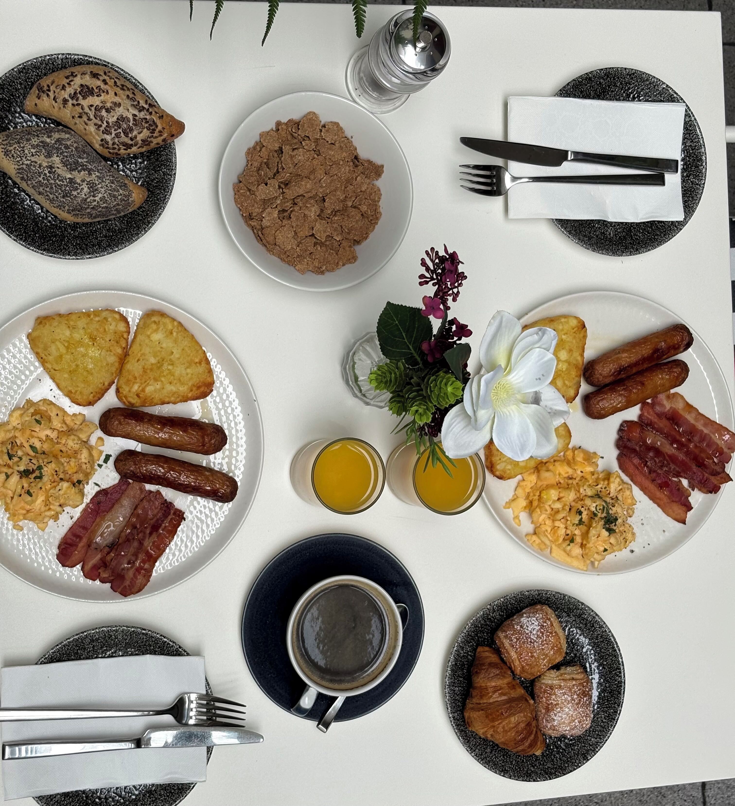 A breakfast setting with plates of eggs, sausages, and pastries, along with a cup of coffee and a flower arrangement.