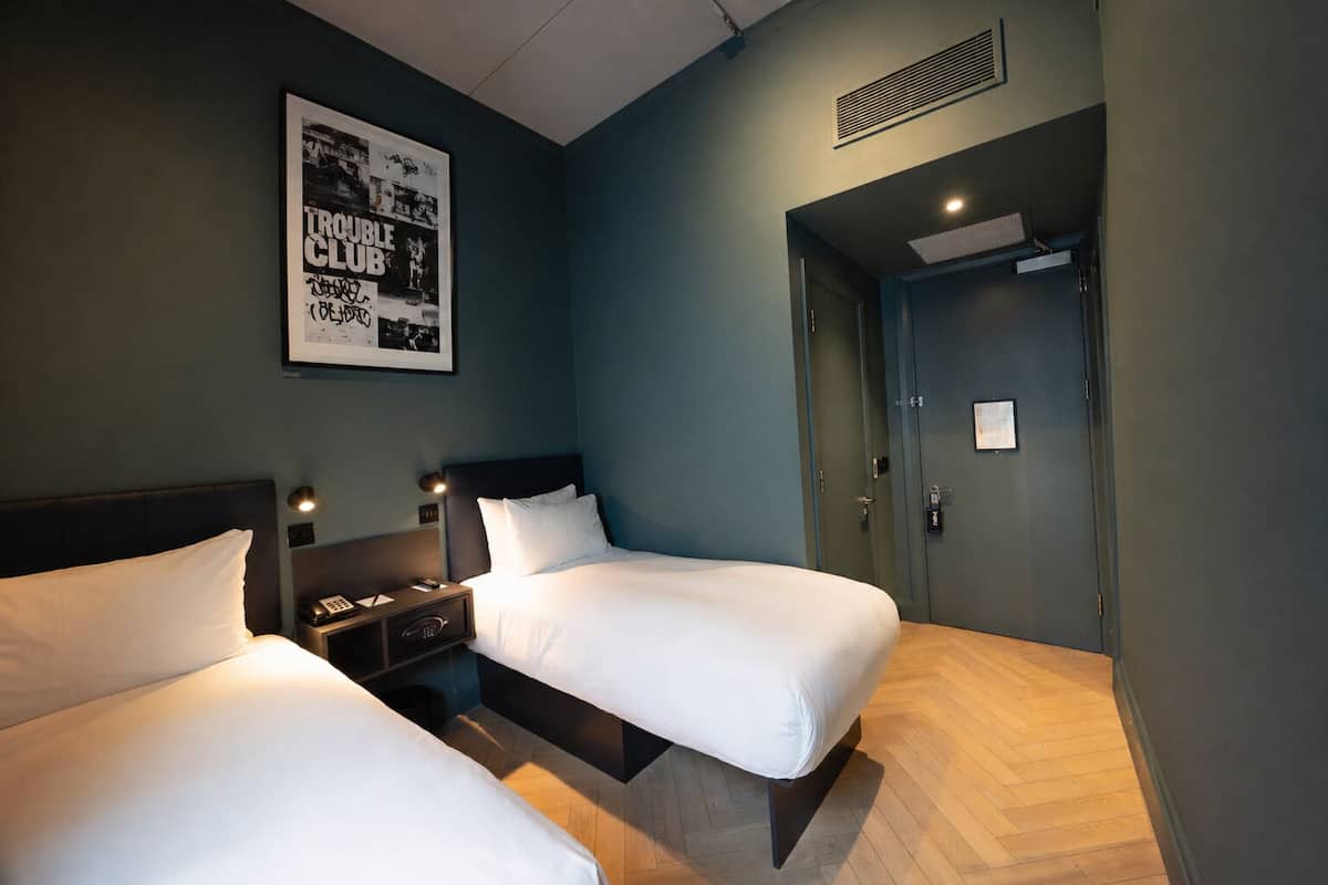 A hotel room with two beds, a nightstand, a wall-mounted poster, and a door with a lock.