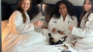 Tres personas vestidas de blanco sentadas en una cama con botellas y copas de champán.