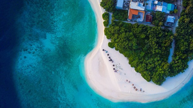 An aerial view of a beach with a row of beach umbrellas and a few people. There is a building with a red roof and several other structures nearby. - Paguro Villa (Ukulhas)