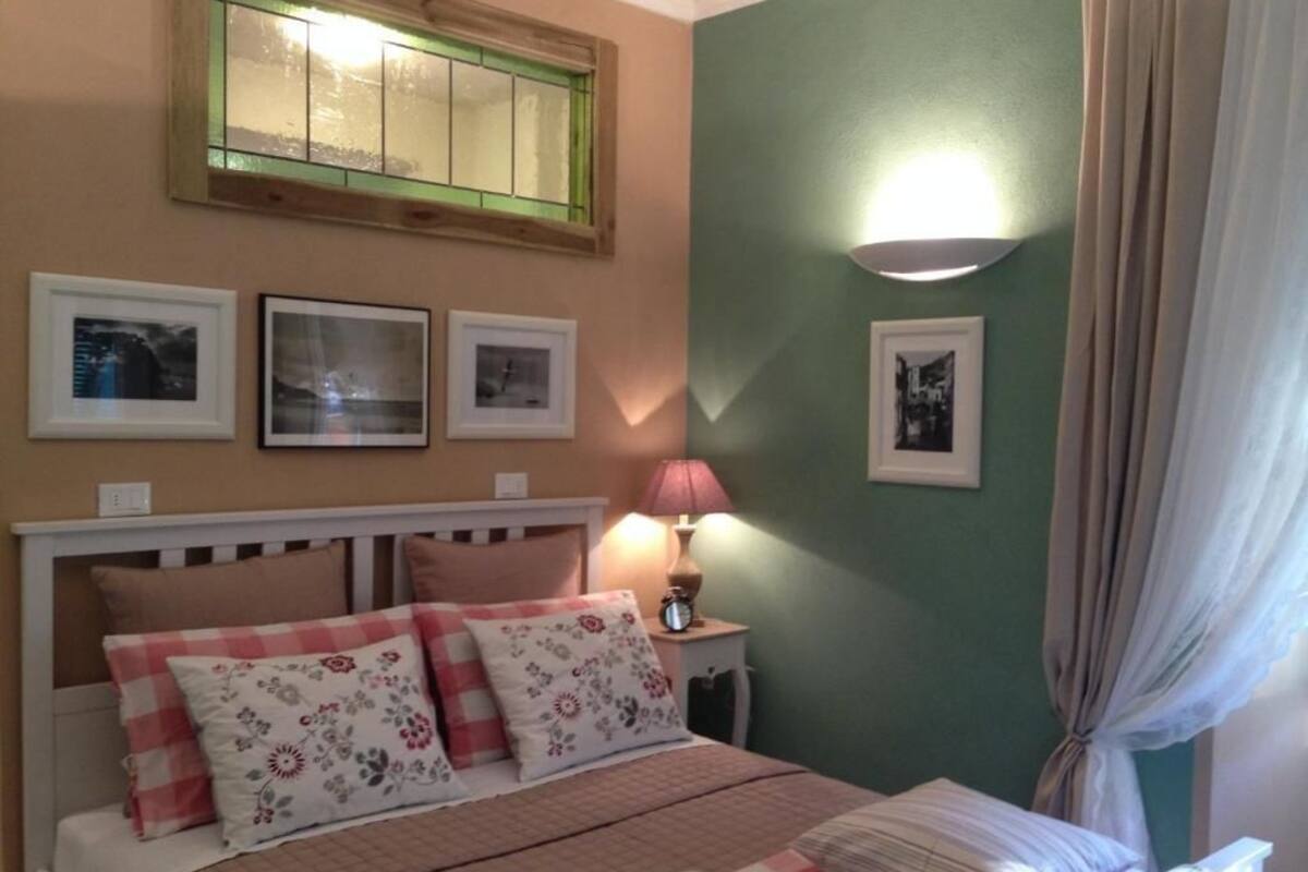 A bedroom with a bed, a nightstand with a lamp, framed pictures on the wall, and a window with curtains.