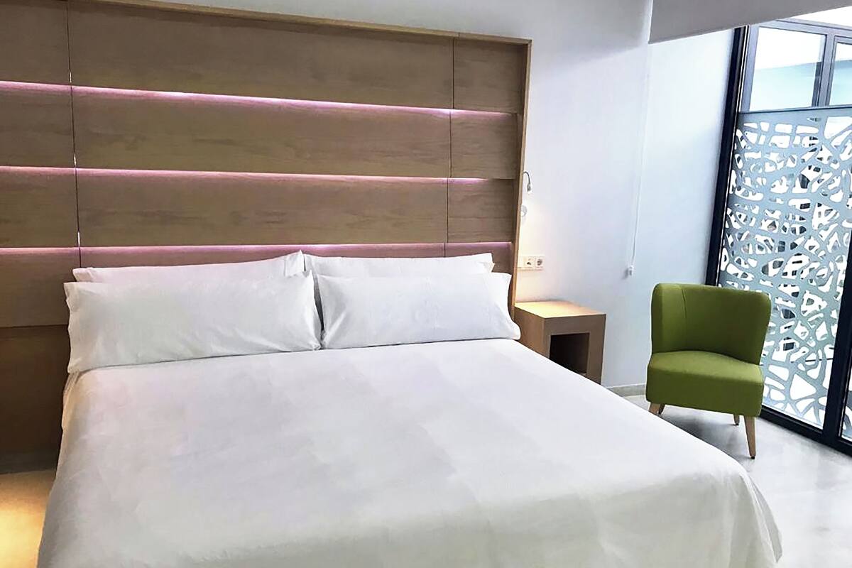 A modern hotel room with a large bed, a wooden headboard, a nightstand, and a green armchair.