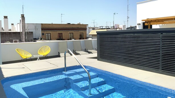 Ein Dachpool mit Stufen, umgeben von einem modernen Gebäude mit schwarzem Rollo und gelben Stühlen.