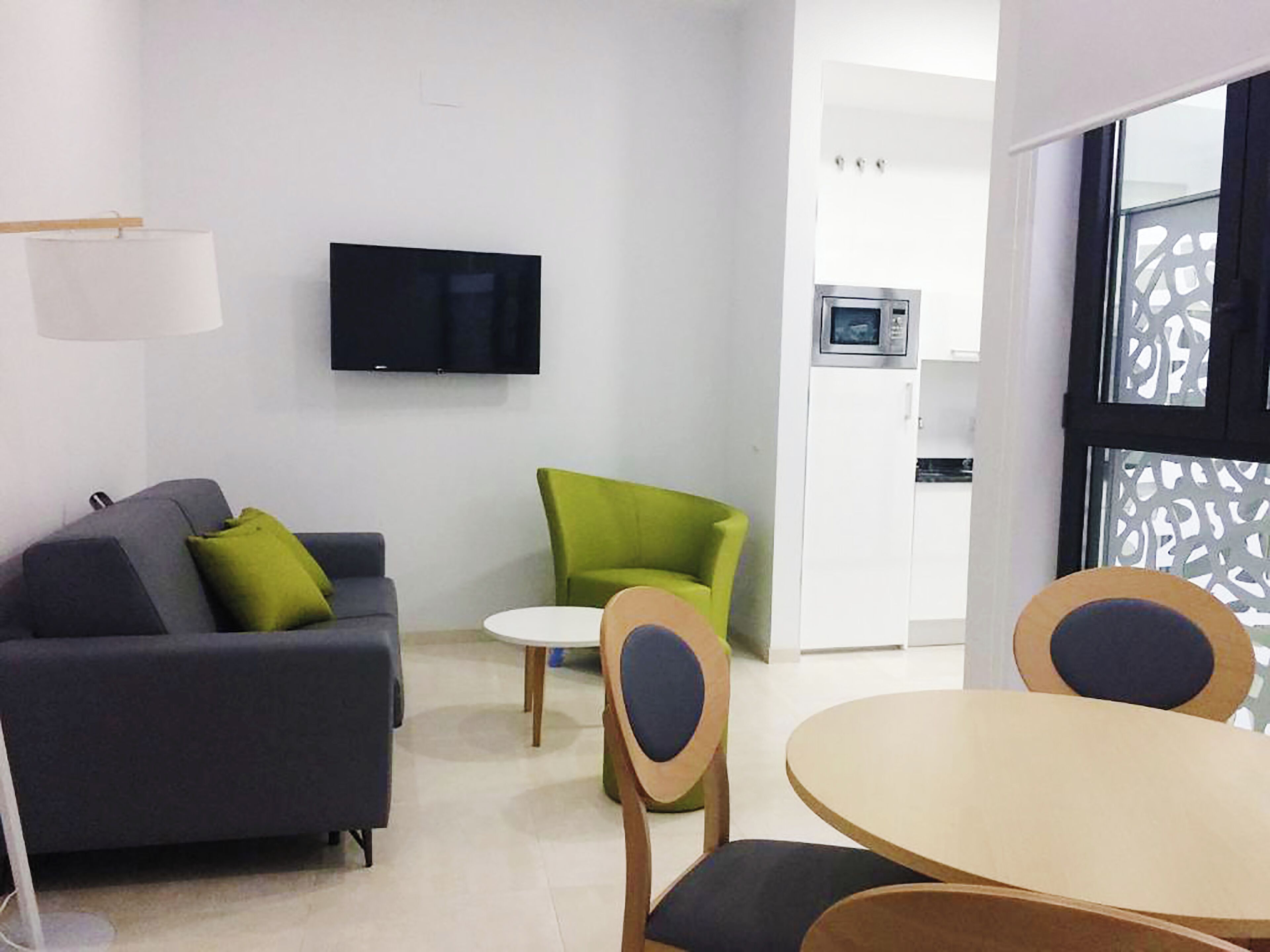 A modern living room with a grey sofa, green armchair, and a round table with wooden chairs. A flat-screen TV is mounted on the wall, and a kitchen area is visible in the background.