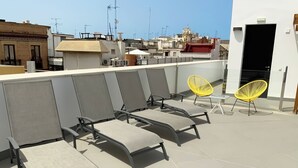 Eine Dachterrasse mit vier grauen Liegestühlen und zwei gelben Sesseln, mit Blick auf eine Stadtlandschaft mit Gebäuden und einer Kirche.