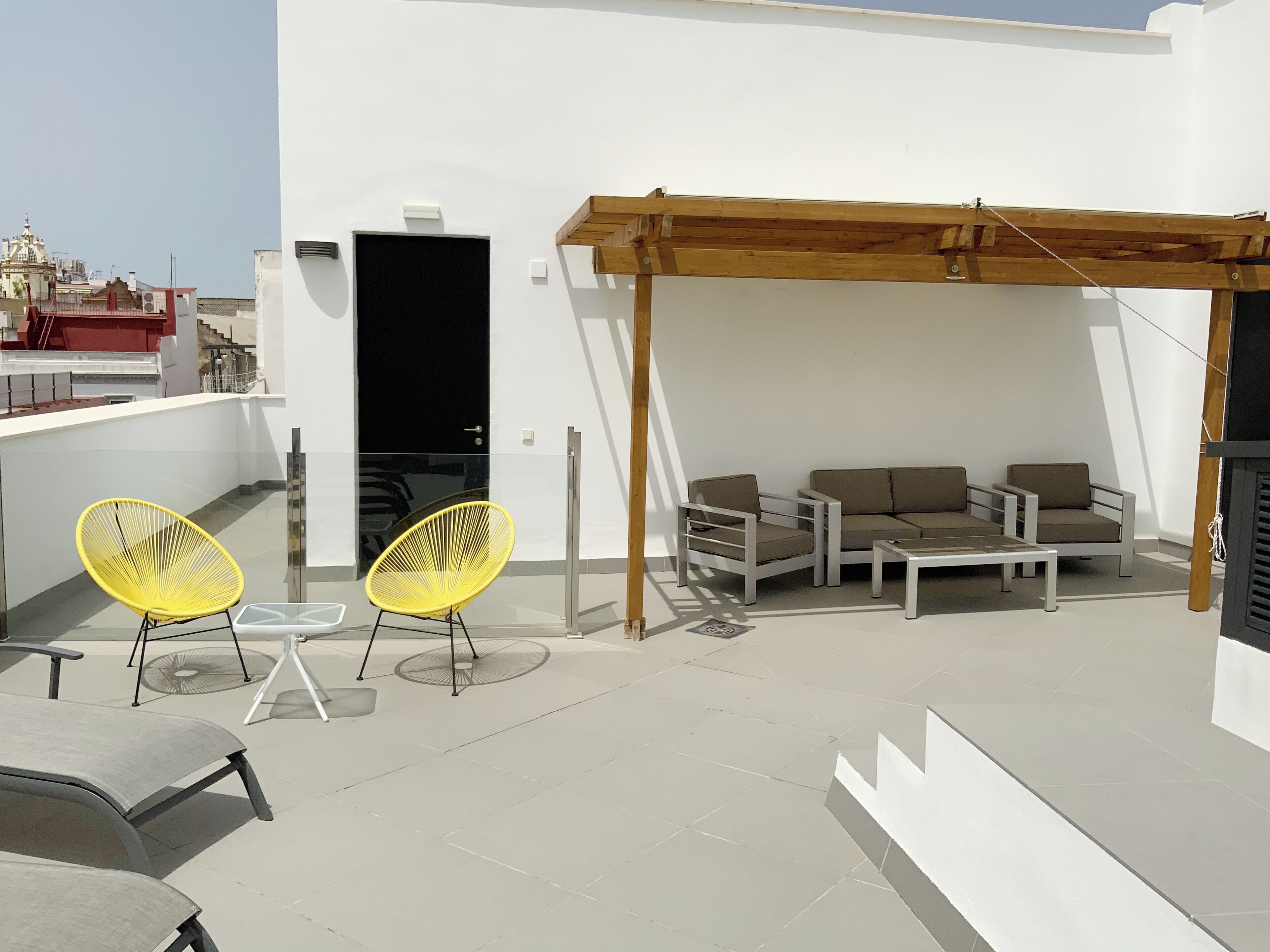 Una terraza en la azotea con una pérgola de madera, dos sillas amarillas, un conjunto de sofás y una mesita.