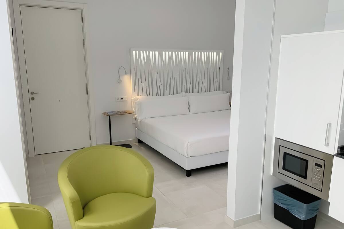 A modern bedroom with a white bed, a green armchair, a small bedside table, and a built-in TV.
