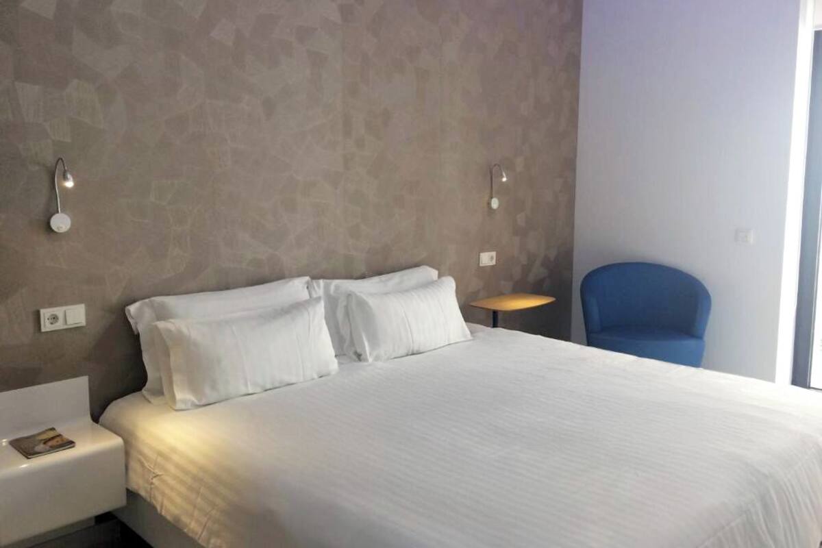 A hotel room with a large bed, two bedside lamps, a small round table, and a blue chair.