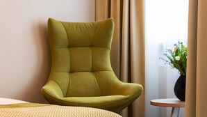 A modern room with a green upholstered armchair, a patterned bedspread, a small round table with a vase of flowers, and a window with curtains. - Gästehaus Lazarus (Berlin)