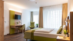 A hotel room with a bed, a desk, a chair, a TV, and a window with curtains. - Gästehaus Lazarus (Berlin)