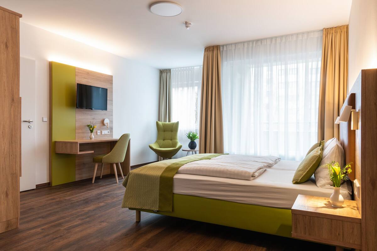 A hotel room with a bed, a desk, a chair, a TV, and a window with curtains.