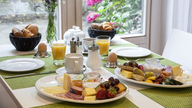 A breakfast table with plates of food, a vase of flowers, and a window view. - Gästehaus Lazarus (Berlin)