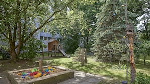 A playground with a slide and climbing structures, surrounded by trees and a wooden playhouse. - Gästehaus Lazarus (Berlin)