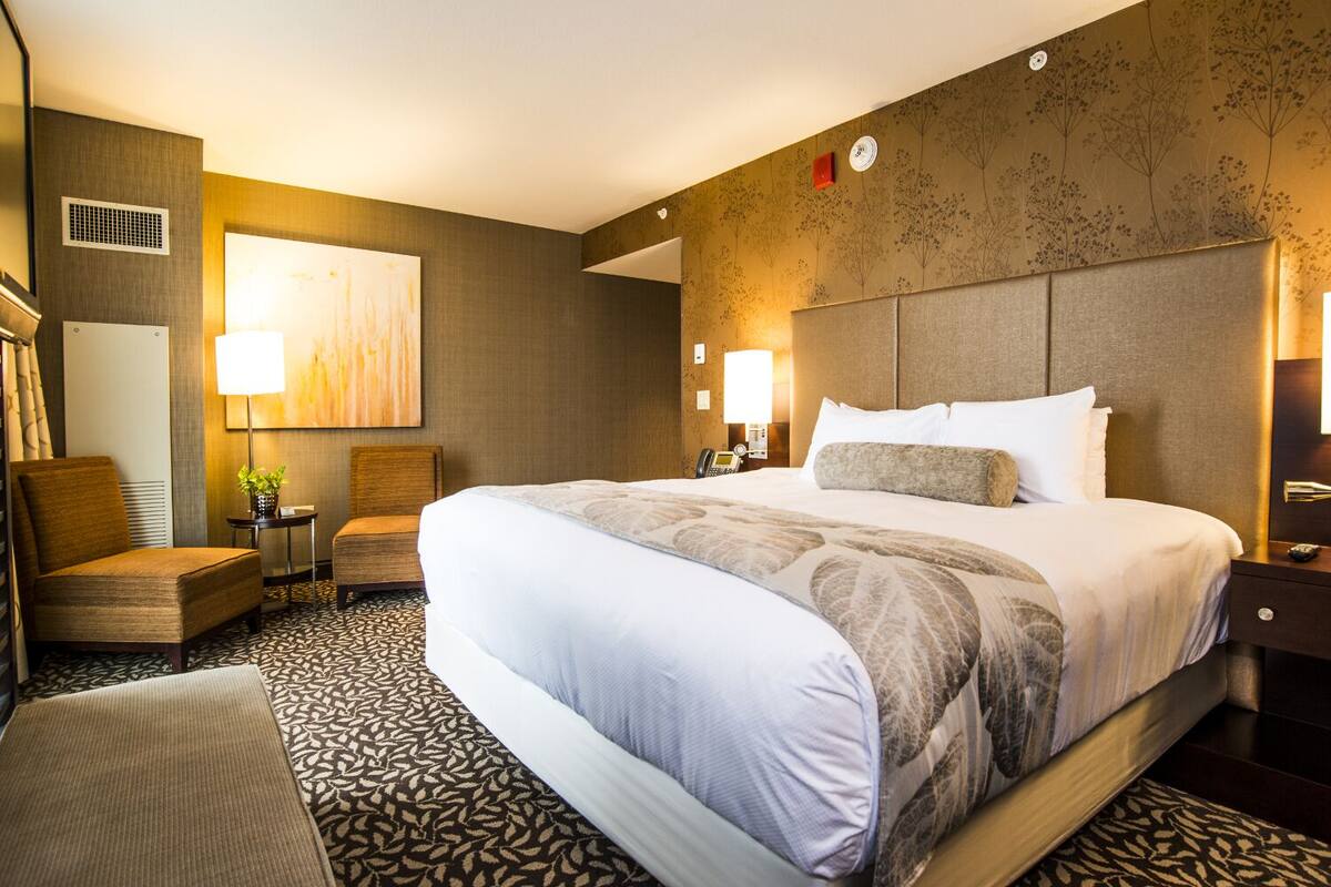 A hotel room with a large bed, two chairs, a desk, and a wall-mounted TV.